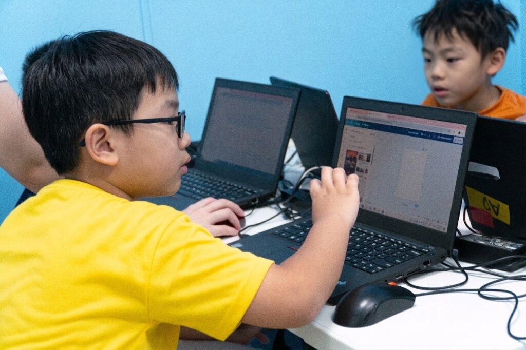 A child working on a laptop during a coding activity, with another child at a nearby laptop.